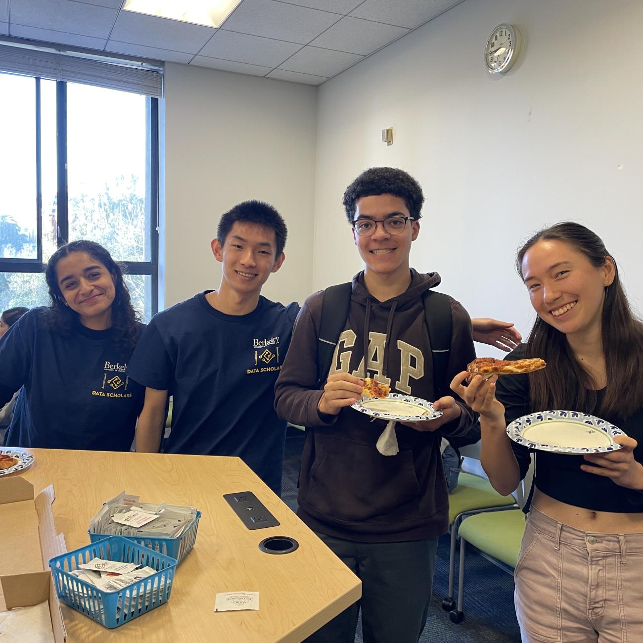 Four Data Scholars students, some holding paper plates with pizza slices, lean in toward each other and smile at the camera