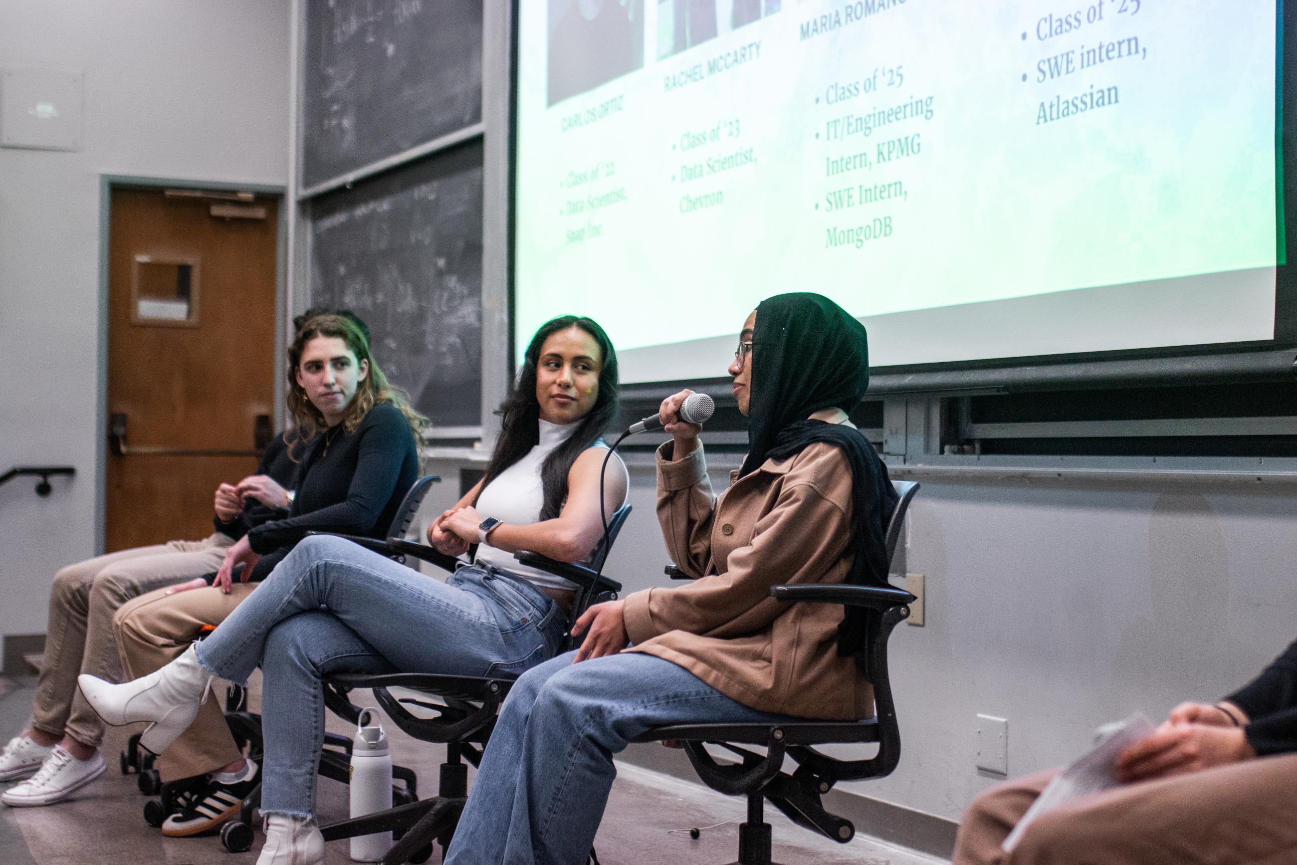 Seated panelist speaks into mic, while other panelists onstage listen.